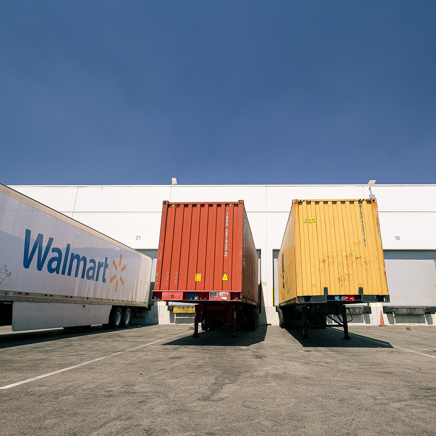 Trucking containers and trailers parked at a logistics facility loading dock. The image shows a white Walmart semi-trailer on the left, and a red and a yellow shipping container backed up to numbered dock doors.