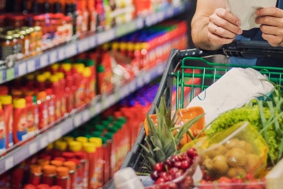 A shopper with a full cart of fresh produce and groceries reviews a shopping list in a store aisle.