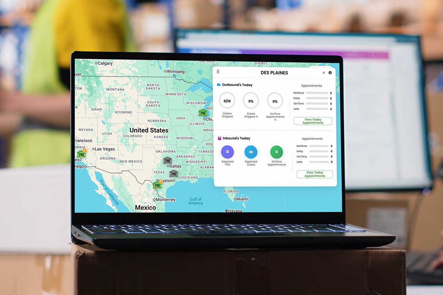Laptop displaying a logistics dashboard with a U.S. map and shipment metrics, placed on a box inside a warehouse with workers and shelving in the background.