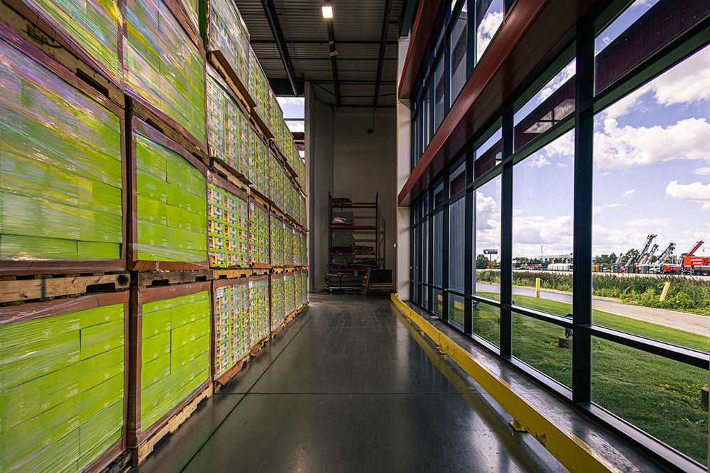 Tall stack of shrink-wrapped lime-green boxes stored along a floor-to-ceiling glass wall in a modern warehouse.