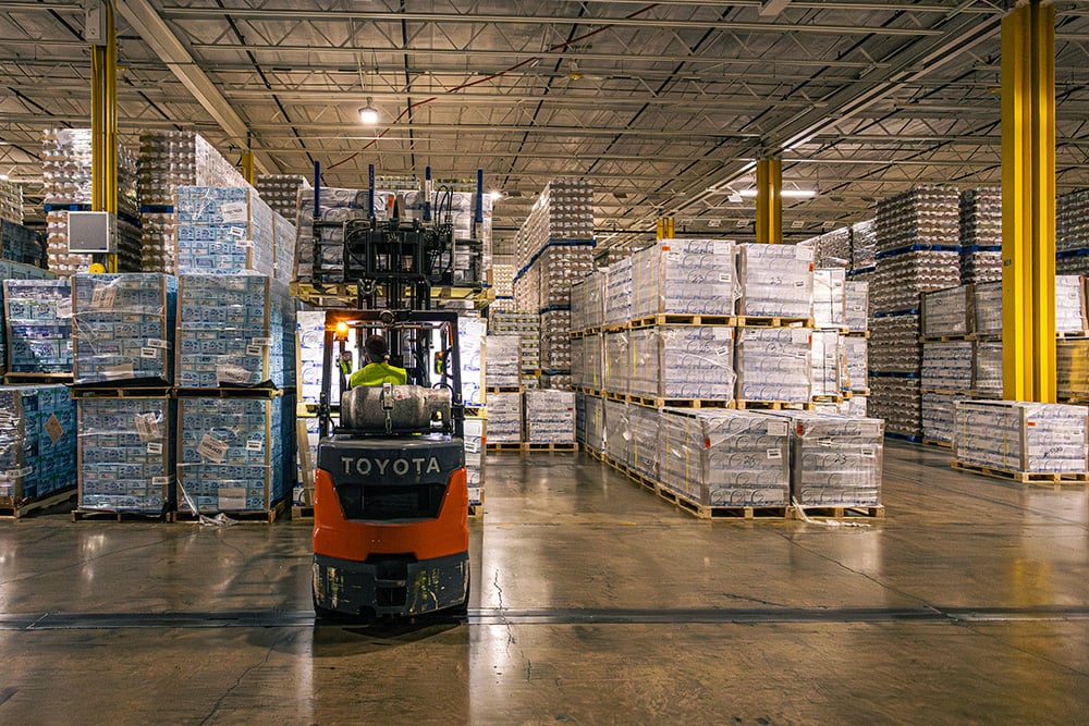 Forklift operator navigating through high-density stacks of shrink-wrapped pallets in a large warehouse.