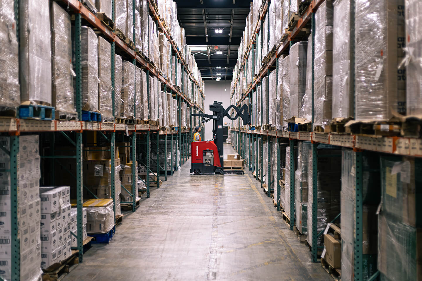 Narrow warehouse aisle with high racks of inventory and a stand-up forklift operating at the end.