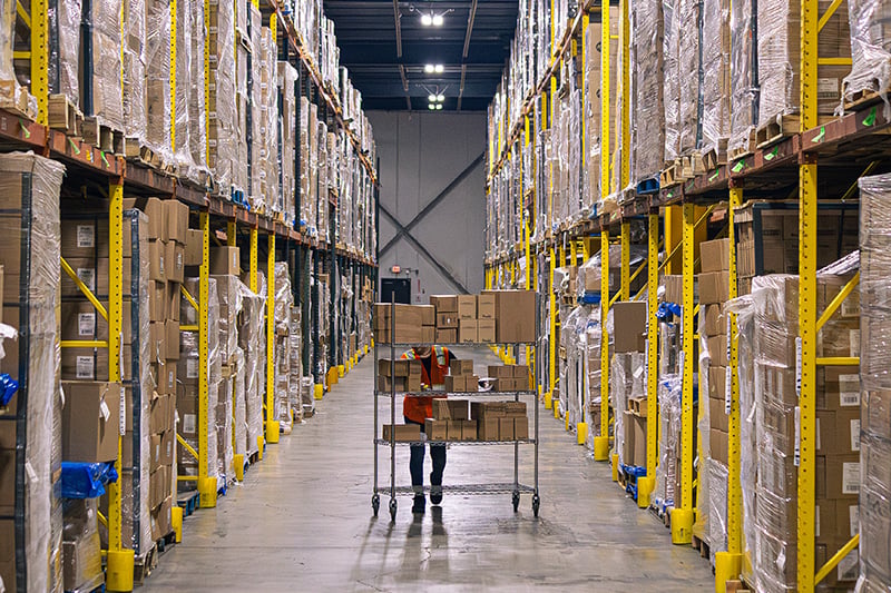 Warehouse worker pushing a wire picking cart through a narrow aisle flanked by tall storage racks.