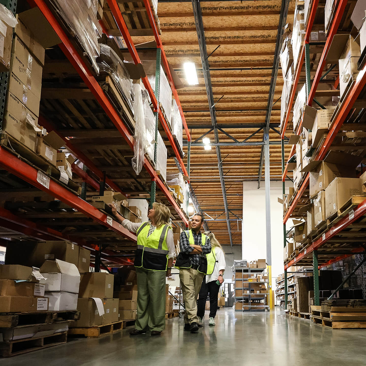 A logistics manager is leading a small team of workers through a warehouse aisle, inspecting items on the red storage racks, demonstrating quality control and inventory management.