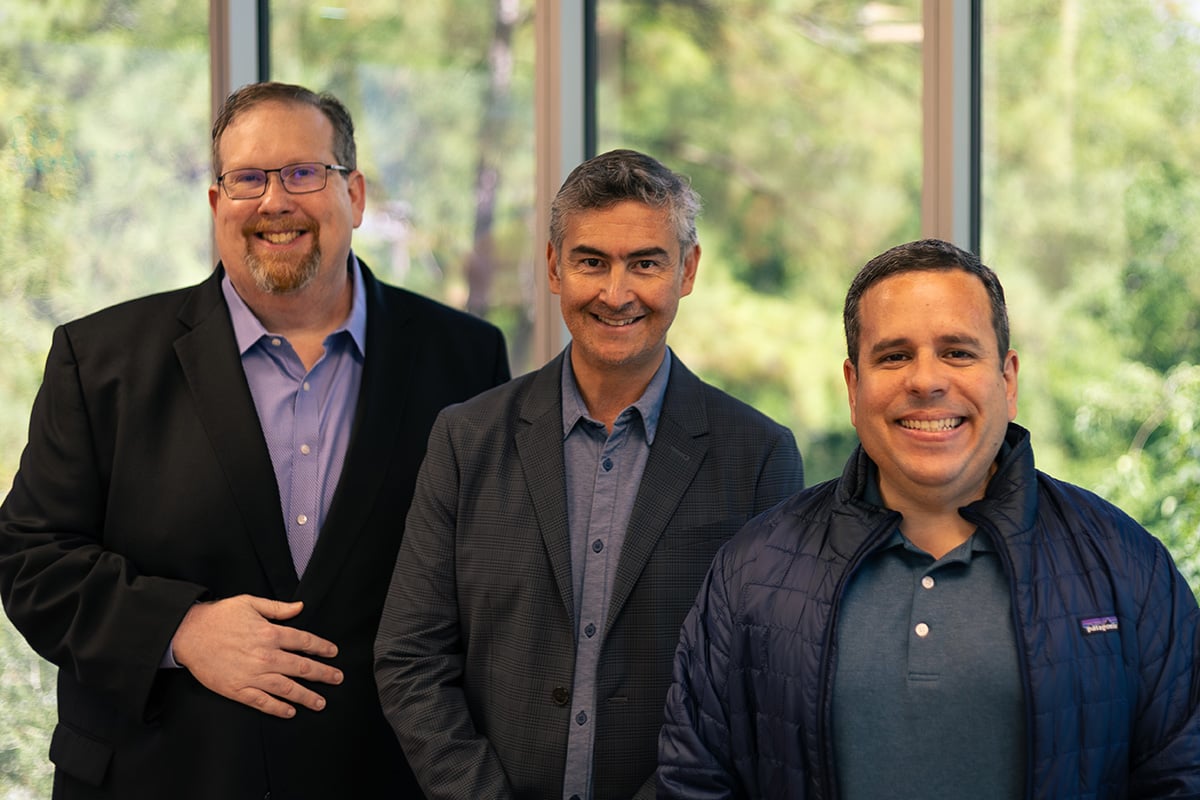 Three male executives from the Source Logistics leadership team pose for a professional portrait in front of a large office window
