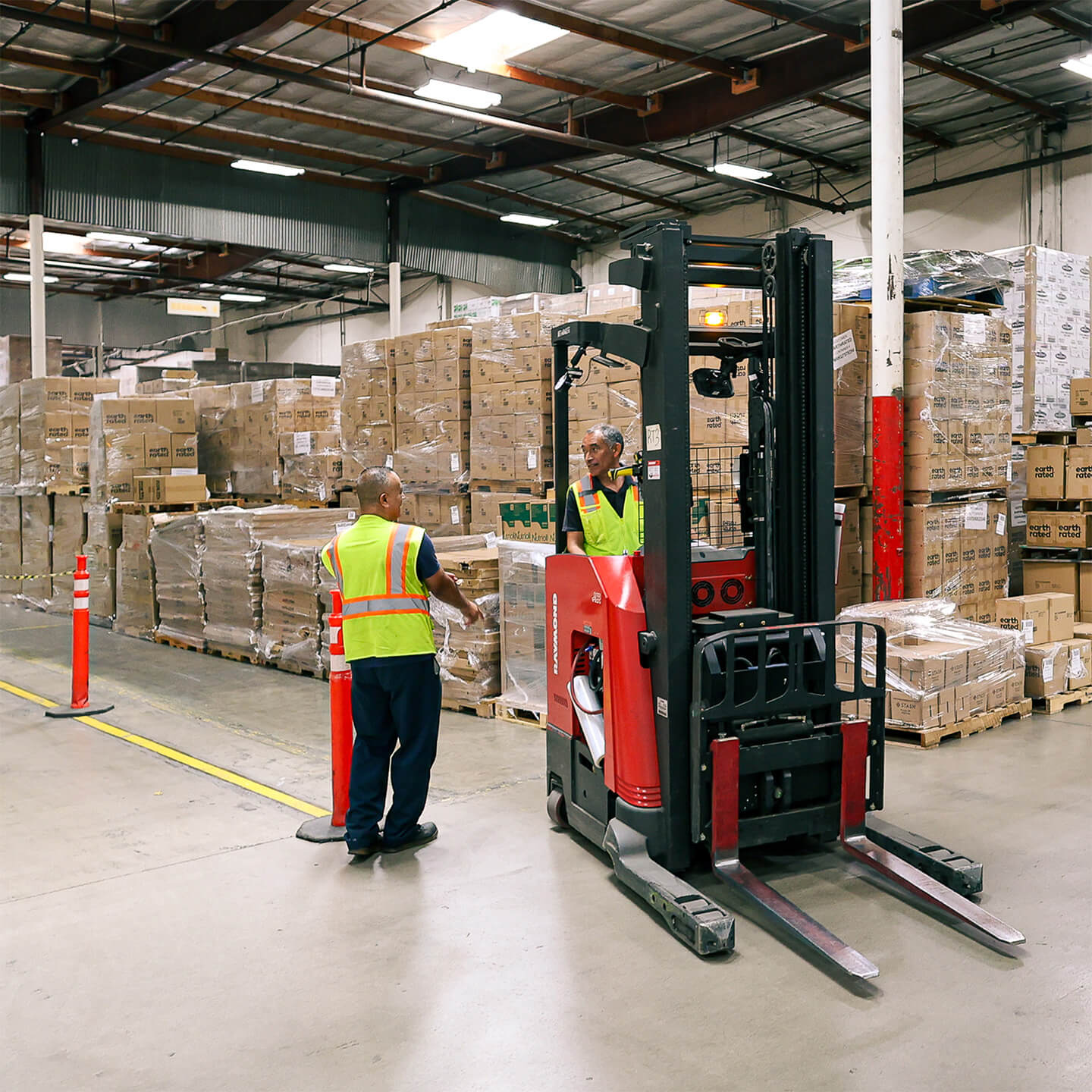 Warehouse workers and a stand-up forklift operating among stacked pallets of inventory.